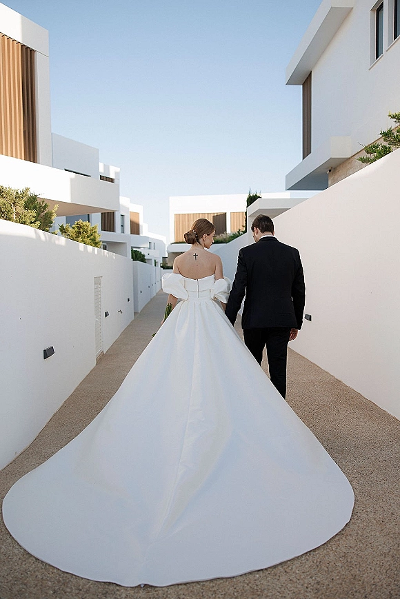 Couple portrait of bride and groom walking away hand in hand, her long train and bouquet flowing on a modern white walkway under blue sky