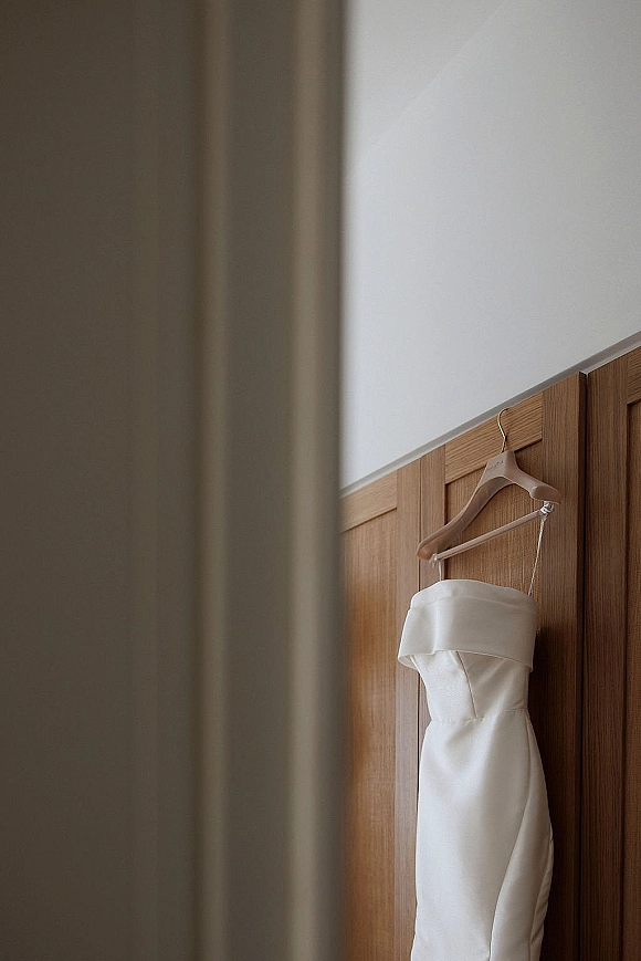 Wedding dress on a wooden hanger, a strapless satin gown with straight neckline hanging against a wood paneled wall and white wall