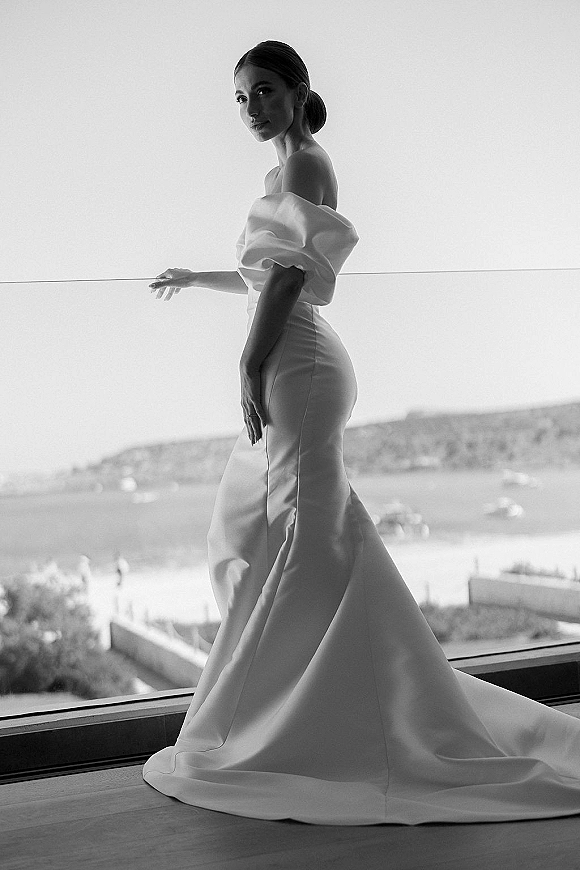 Bridal portrait of a bride in a wedding dress with train, off the shoulder satin gown, by a large window with ocean views and boats