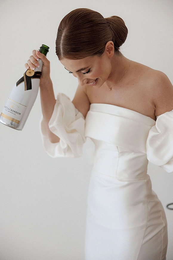 Bridal portrait of a smiling bride holding champagne bottle with a ribbon bow, wearing an off-the-shoulder puff-sleeve gown by a neutral wall