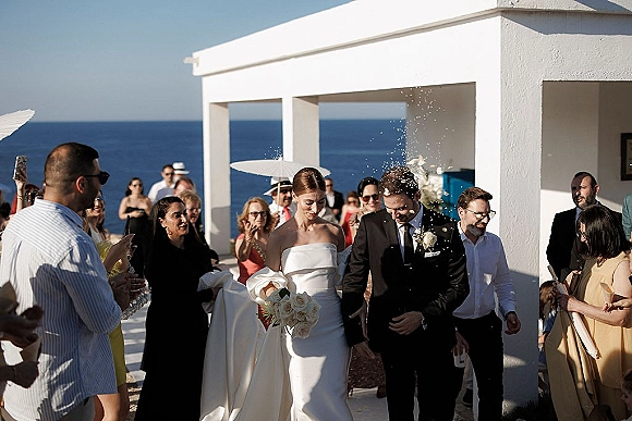 Wedding recessional as bride and groom walk the aisle through cheering guests, confetti falling on an ocean-view terrace by a white venue