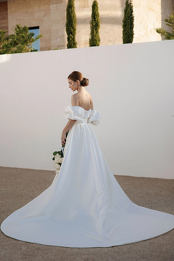 Bridal portrait of a bride in an off the shoulder wedding dress holding a bouquet, standing by a white wall in a courtyard with cypress trees