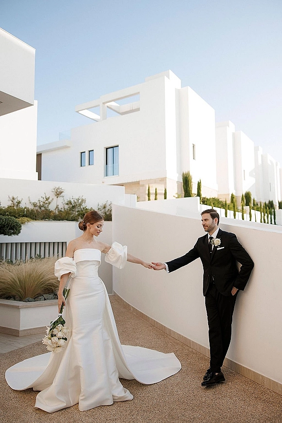Couple portrait of bride and groom holding hands, her puff-sleeved strapless gown with long train and white rose bouquet on modern terrace