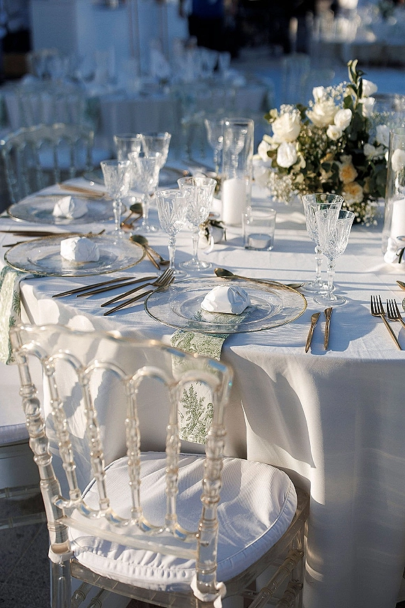 Reception tablescape with a white wedding table setting featuring crystal goblets, gold flatware, white flowers, and pillar candles outdoors in soft sunlight