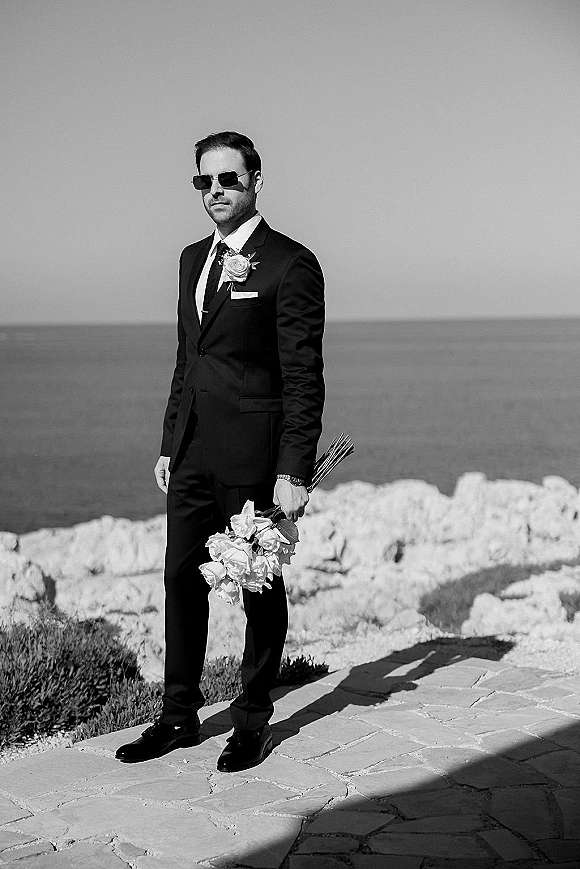 Groom portrait in a black suit wearing sunglasses, holding a bouquet on a stone patio with ocean horizon and rocky shoreline behind