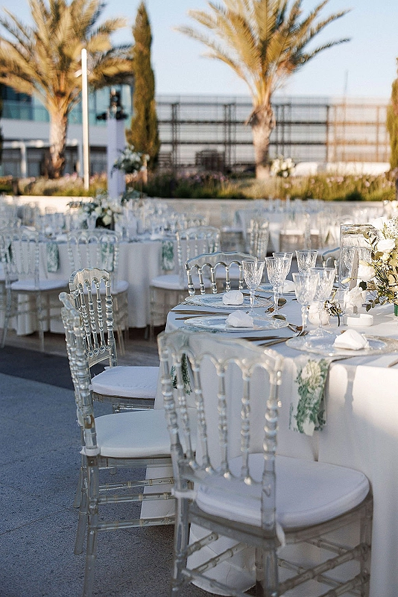Reception tablescape with white wedding table decor, clear acrylic chairs, glass goblets, floral centerpiece, and votive candles on a terrace with palms