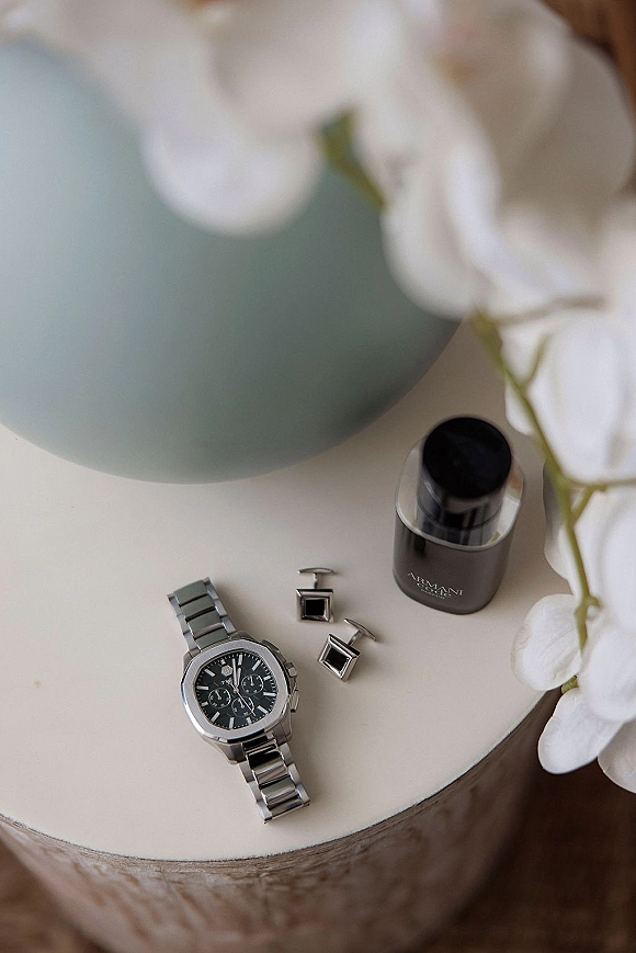 Groom accessories flatlay with a metal wristwatch, cufflinks and cologne bottle beside white flowers on a round side table over wood floor