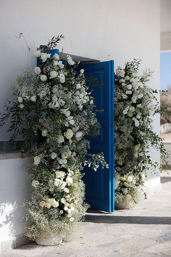 Wedding floral arch framing a blue door with white roses and baby's breath, lush greenery, and floral pillars in a stucco courtyard