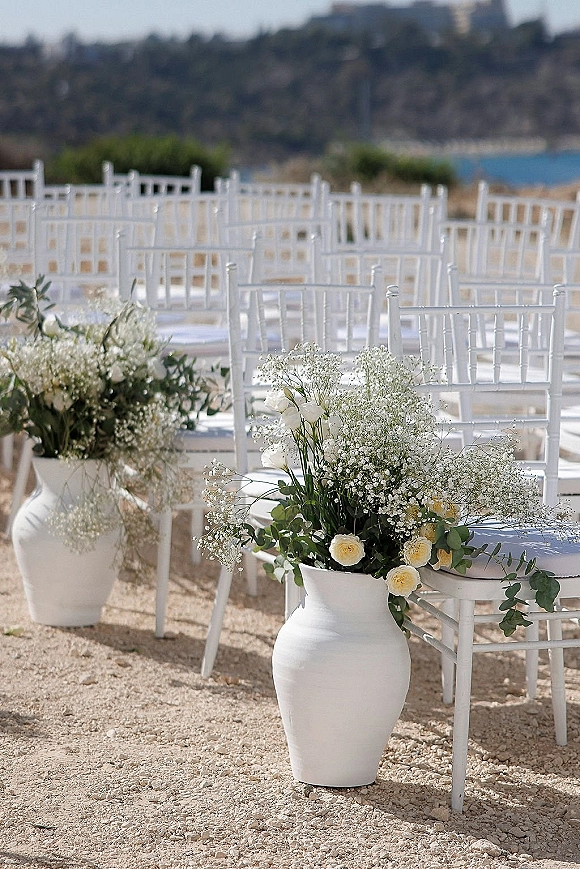 Ceremony setup with outdoor ceremony chairs lining a gravel aisle, white vases of roses and baby's breath beside water and hills beyond
