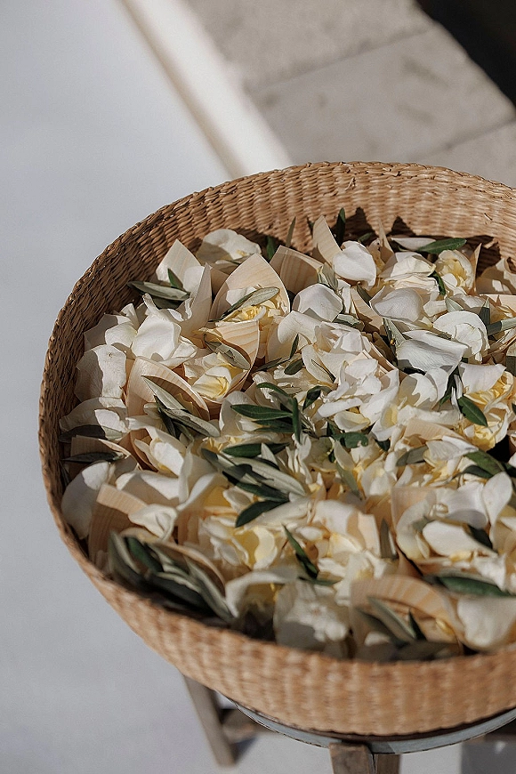 Wedding petal toss setup with white flower petals and greenery in a woven basket and wooden cones on a stone floor by a white wall