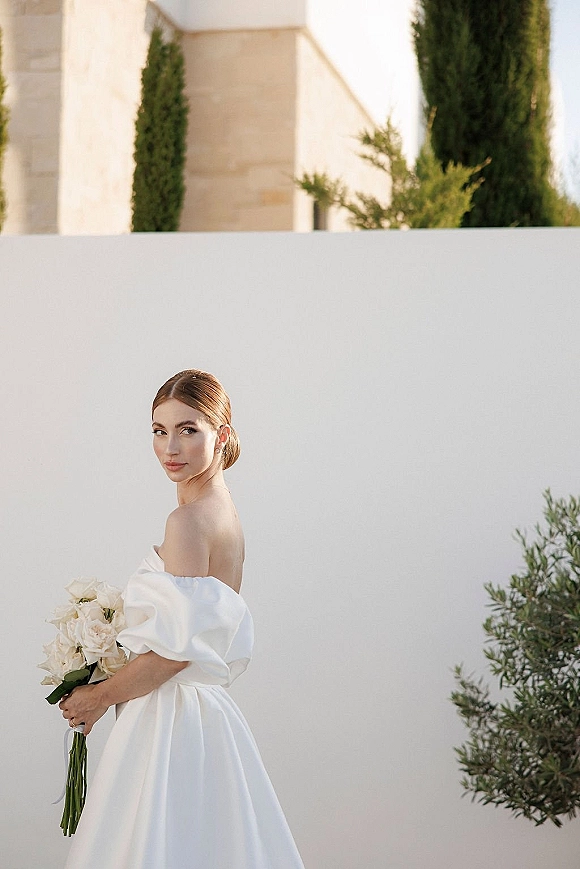 Bridal portrait of a bride in an off the shoulder wedding dress holding a white rose bouquet, posed by a white wall under blue sky