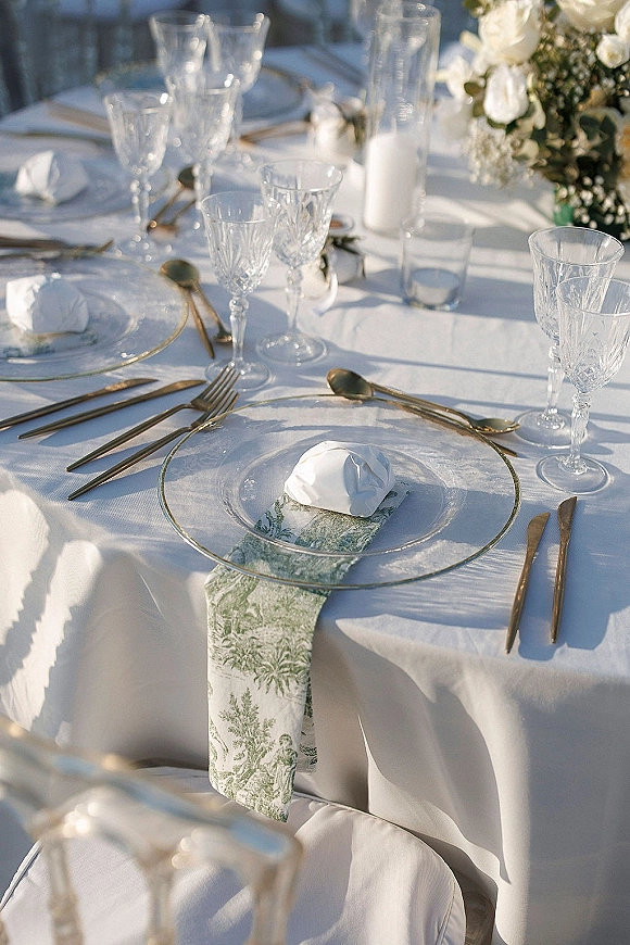 Reception tablescape with wedding place setting, clear glass charger plates, crystal stemware, gold flatware, and tall cylinder candle in outdoor light