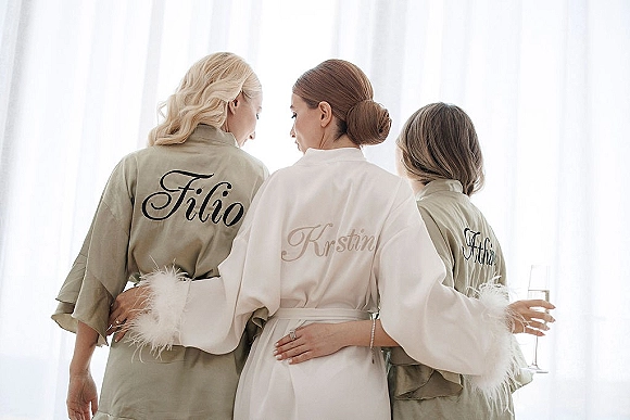Bridesmaids robes in personalized satin with monogram embroidery, shown in window light by sheer white curtains, champagne flute nearby