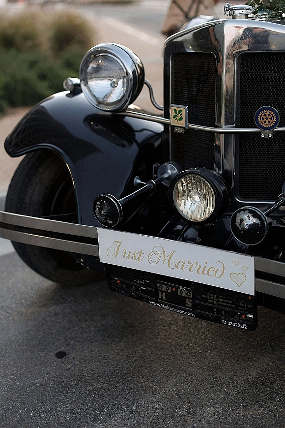 Just married sign with calligraphy lettering and heart doodles on a vintage car grille and chrome headlights, parked on a road with greenery