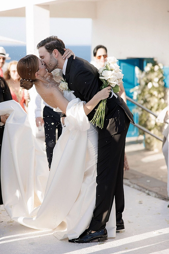Wedding kiss as bride in veil and gown holds white bouquet while groom in tuxedo dips her amid confetti on seaside terrace