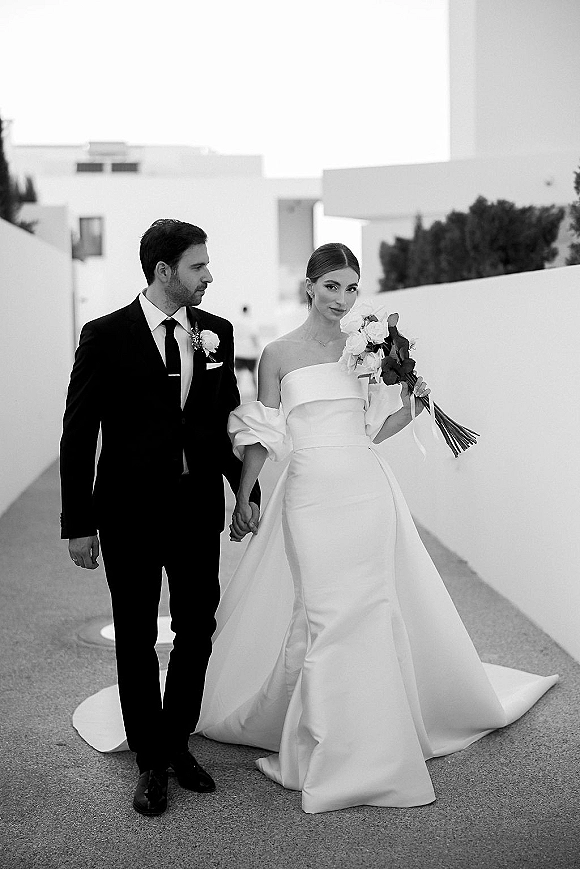 Couple portrait in a black and white wedding photo, bride in strapless gown with train holding bouquet as groom holds her hand on walkway