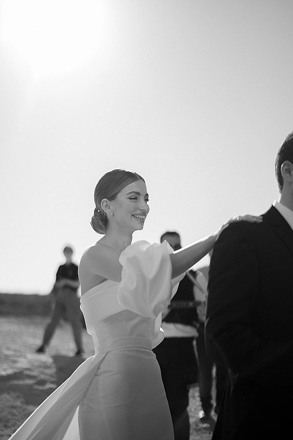 Bride and groom dancing during a first dance outdoors, bride in strapless dress with waist bow as guests watch by the shoreline sky
