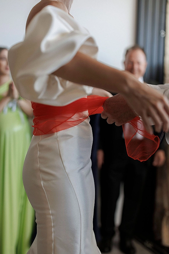Wedding reception dance as bride in a white dress twirls with a red tulle sash while partner in suit jacket holds hands, guests watch in window light