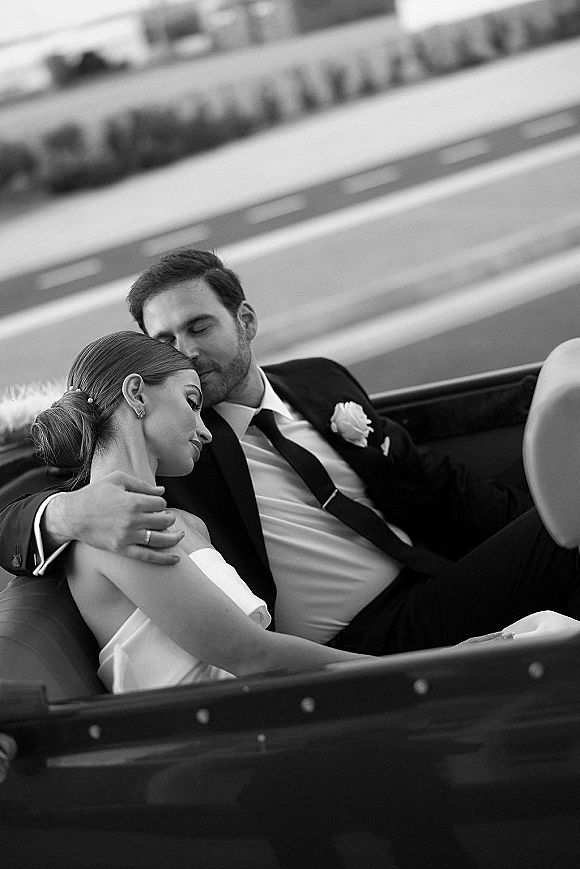 Couple portrait in black and white wedding portrait style, bride resting on groom’s shoulder in a convertible on a city street, earrings glinting