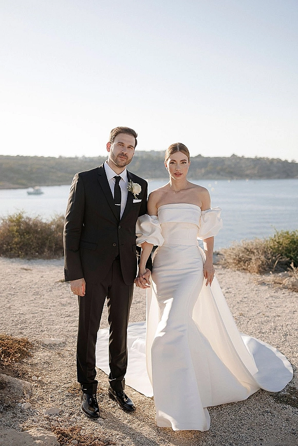 Couple portrait of bride and groom holding hands, full length on a coastal path with ocean cliffs behind, her dress train flowing