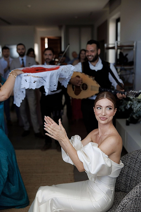 Bride getting ready clapping in a strapless satin wedding dress, smiling as a musician plays in a window-lit hotel room with guests