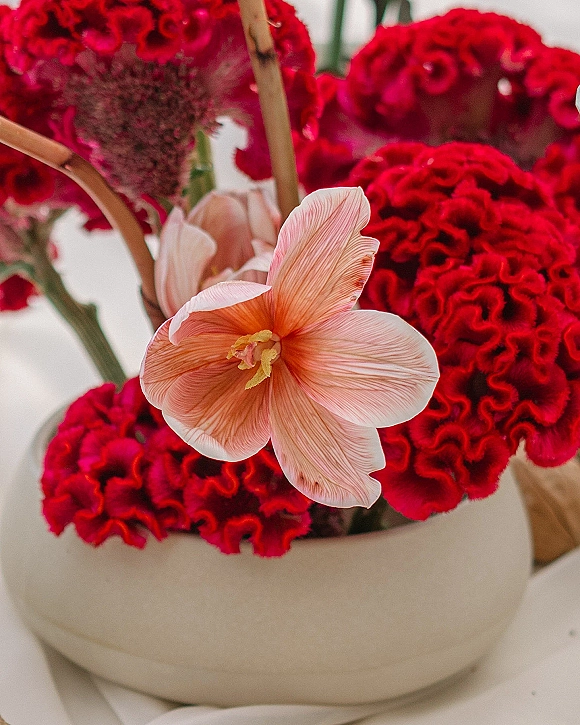 Wedding centerpiece flowers in a ceramic bowl featuring red celosia and pink anthurium stems on a white tabletop against a neutral backdrop