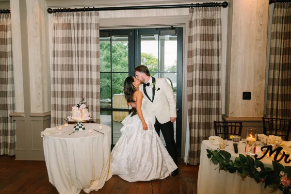 Wedding kiss portrait of the couple sharing a cake cutting kiss beside their wedding cake, framed by a window and distant trees.