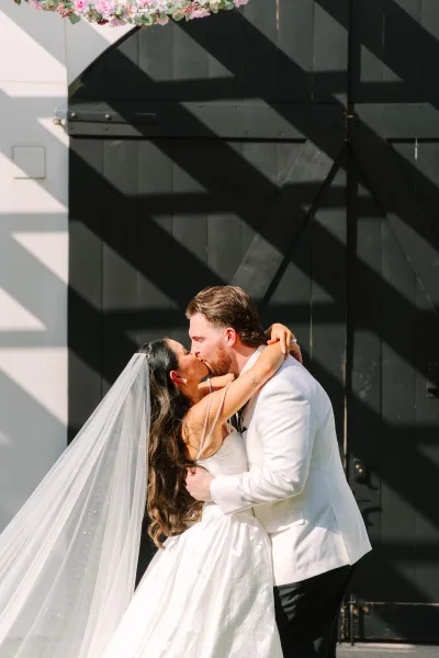 Wedding kiss portrait of a bride and groom embrace, highlighted by a pearl shoulder drape before black barn doors with sunlit shadows.