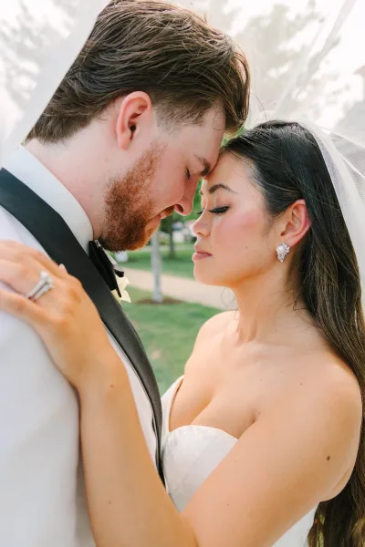 Couple portrait of a bride and groom embrace, foreheads touching as her bridal veil softly frames them, her hand on his lapel in natural light.