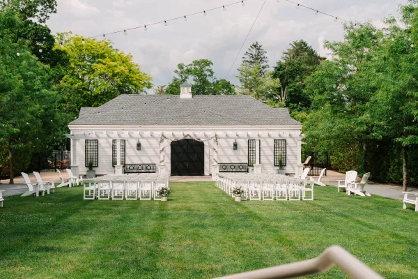 Outdoor ceremony setup with thoughtful ceremony aisle design, white chairs arranged symmetrically on a lawn before a white building.