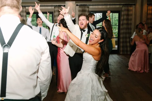 Wedding reception dance with the bride and groom dancing amid cheering guests, his white tux jacket sharp against wood floors and tall windows.