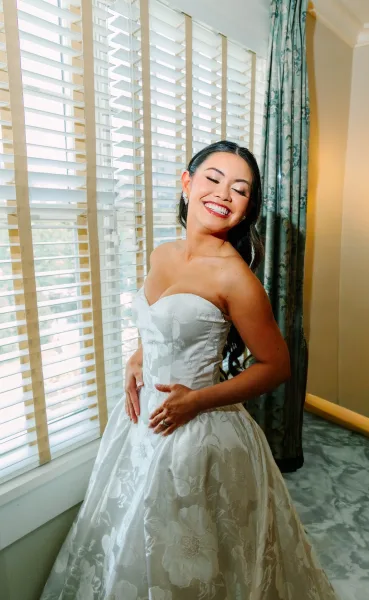 Bridal portrait of a bride in strapless gown with a sweetheart neckline and textured floral skirt, smiling with hands resting at her waist.
