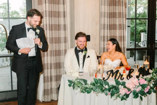 Wedding toast captures a reception speech moment as a friend speaks into a microphone while the bride laughs and the groom smiles.