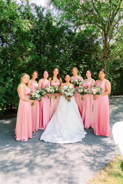 Bride and bridesmaids in pink bridesmaid dresses pose on a gravel path under garden trees, framed by a lawn.