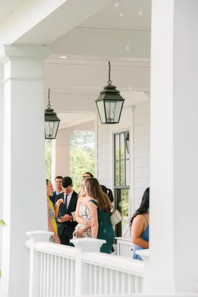Wedding guests in a guest arrival moment mingle on a white-columned porch, lanterns hanging, with garden trees beyond and windows behind.