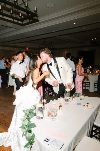 Wedding kiss moment as they share a sweetheart table kiss, framed by floating candles, with guests dancing under ballroom chandeliers.