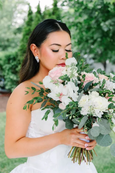 Bridal portrait of a bride with bouquet in a strapless bridal gown, amid garden greenery and trees, softly lit by natural light.