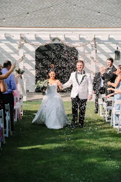Wedding exit during a petal toss exit as newlyweds hold hands, the bride lifting a white-and-green bouquet by a white building's arched doorway.