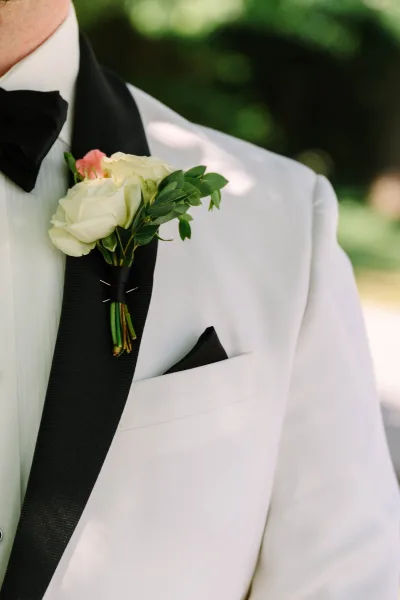 Groom boutonniere, a white rose boutonniere with a pink bud detail, set against garden greenery in the background.