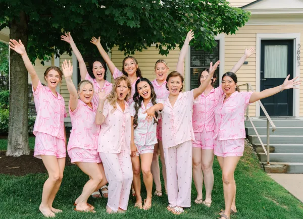 Bridal party portrait with bridesmaids in pajamas cheering on house steps, wearing pink pajama sets and sandals, tree and lawn in the background