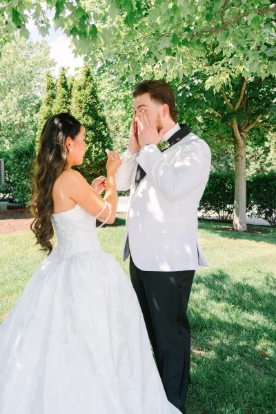 Wedding first look with a groom crying as the bride offers a handkerchief in sun-dappled garden light beneath trees and hedges.