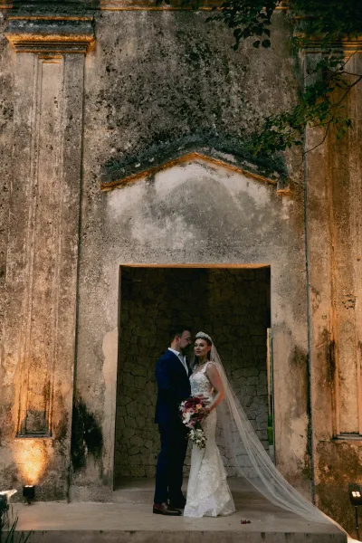 Couple portrait of bride in lace gown with bridal tiara and veil as groom kisses her forehead by an ivy stone archway with uplighting