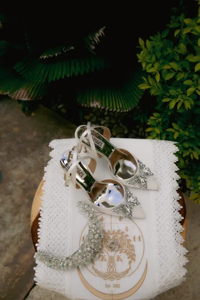 Bridal shoes white wedding heels with crystal bow clips on a lace runner beside pearl jewelry and invitation, greenery walkway behind