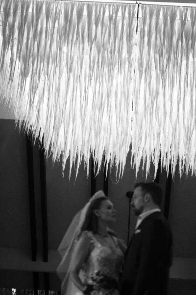 Ceremony moment as bride and groom at altar stand under ceiling drapery and hanging fringe, her veil and gown set against dark beams backdrop
