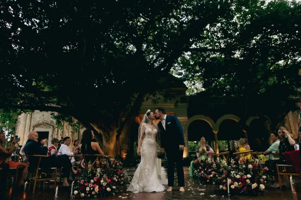 Ceremony kiss as bride in veil and tiara kisses groom in navy suit on a rose petal aisle under a tree canopy in an arched courtyard