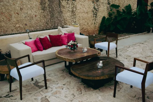 Wedding lounge seating with a white sofa and pink throw pillows, wooden chairs, and candlelit floral tables on a stone patio by greenery
