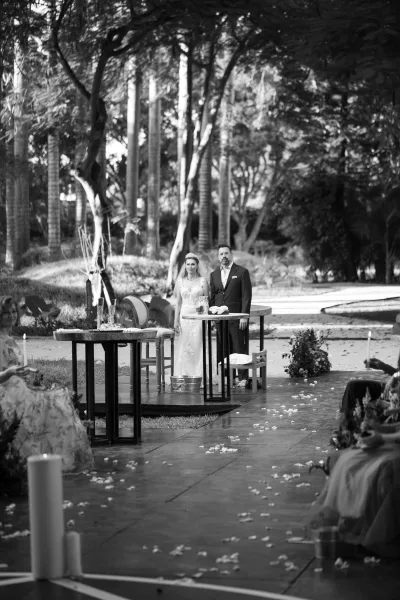 Ceremony moment as bride in veil and tiara faces groom in dark suit on a stone aisle with flower petals and candles in a garden