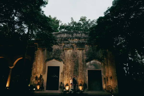 Couple portrait of bride in lace dress and veil holding a red bouquet beside groom in black suit, lit at night before an arched historic doorway