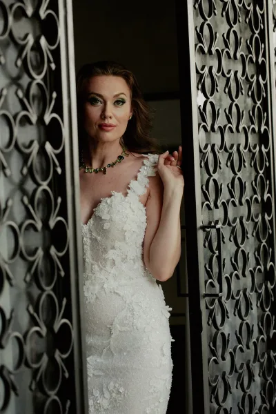 Bridal portrait of a bride in a lace, beaded gown with emerald necklace, standing in a doorway by a wrought iron gate in dim interior