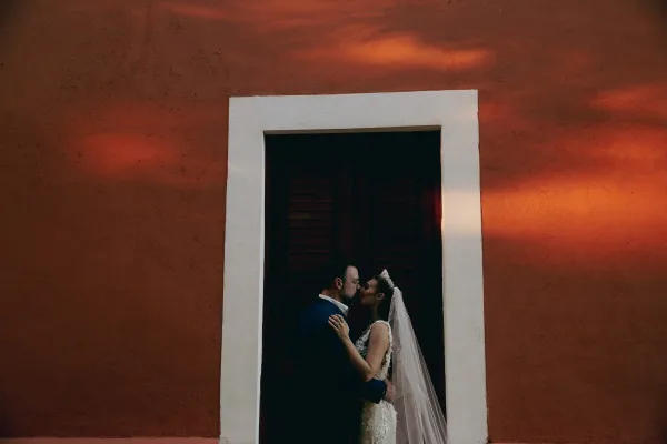 Wedding kiss portrait of bride and groom kissing, her veil and tiara framing a lace dress, by a sunlit stucco doorway with shadows